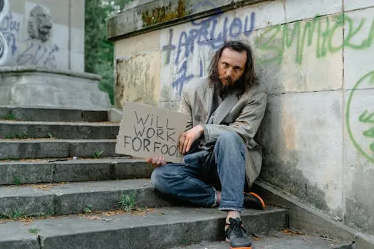 A homeless man sits on steps outdoors holding a 'Will Work for Food' sign, depicting poverty.